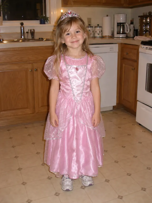 Young girl in a pink satin dress-up princess costume and toy tiara standing in a kitchen